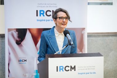 Viviane Poupon se tient derrière un lutrin portant le logo « IRCM », s’adressant à un micro lors d’un événement intérieur. Une bannière avec l’inscription « IRCM » et un visuel scientifique est visible en arrière-plan.