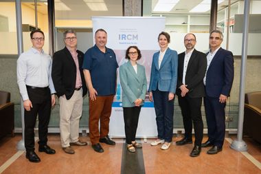 A group of seven professionals stands in a hallway or office corridor, positioned in front of a poster on an easel displaying the text “IRCM.” The individuals are arranged in a line, wearing business or business-casual attire in neutral and blue tones. The setting includes glass walls, beige flooring, and a well-lit interior, suggesting a research environment.