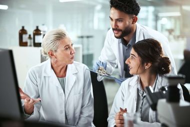 Three researchers in lab coats discussing work together in a laboratory, with one holding a clipboard near a glass equipment cabinet.