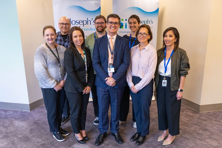 Dr. Benicio Frey stands at the centre of a group of nine colleagues posing in two rows in a hospital lobby. They are smiling in front of St. Joseph's Healthcare Hamilton banners, and most wear business attire and lanyards with ID badges.